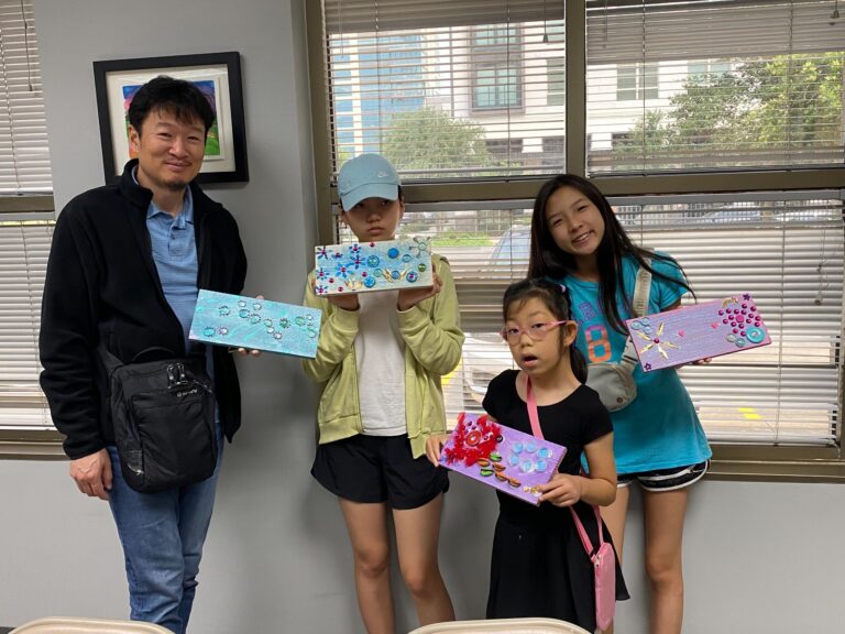 Three artists and a volunteer hold up their finished boards side by side, each decorated with a different arrangement of gems, buttons and painted flowers.