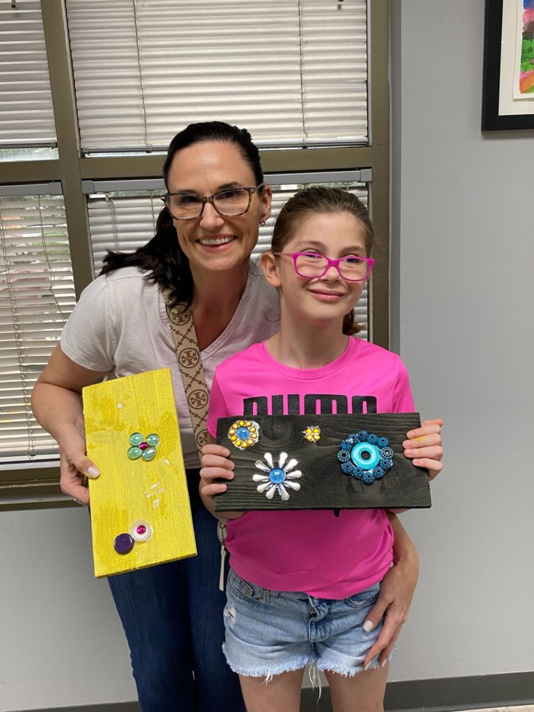 An artist and a volunteer hold up two finished boards together: one painted yellow with gem flowers, the other painted dark with layered flower shapes in blue, white and yellow.