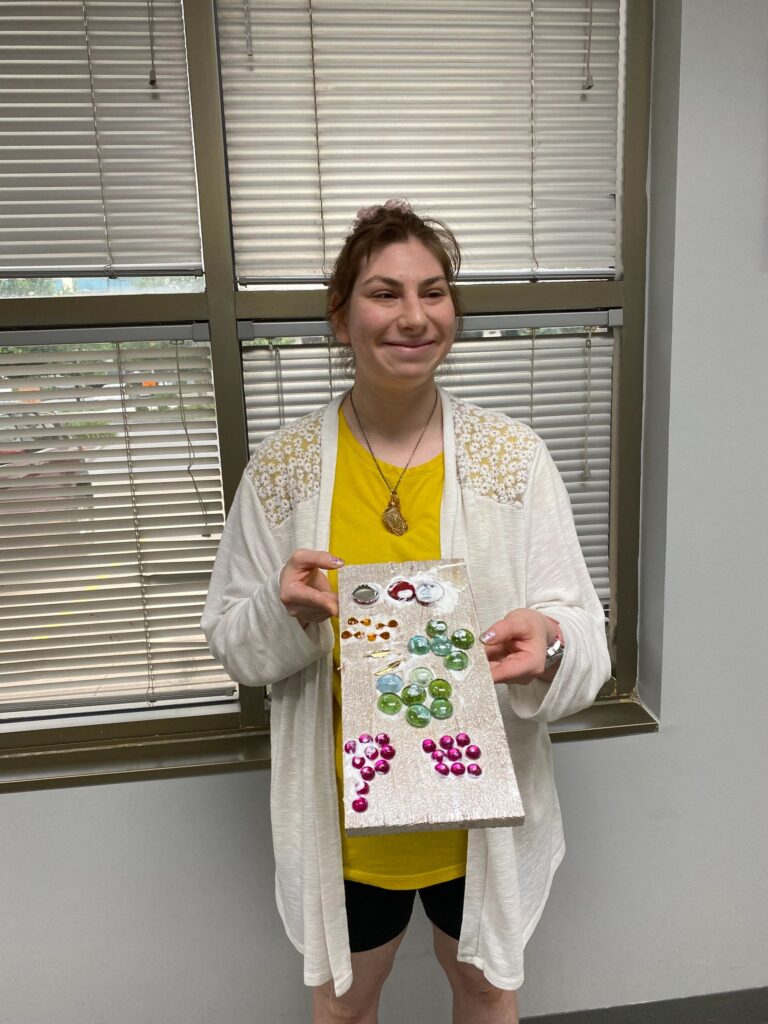 An artist holds a finished board painted cream and decorated with clusters of glass gems in red, green and pink.