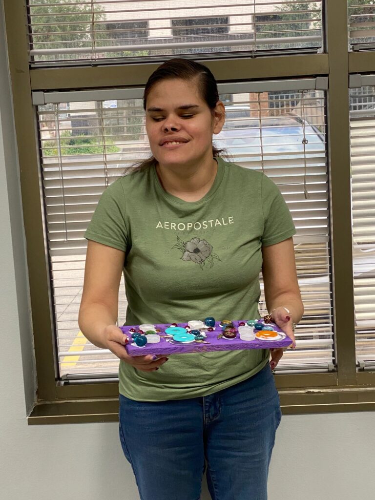 An artist holds a finished board painted purple and decorated with a cluster of glass gems and buttons in teal, pink and cream near one end.