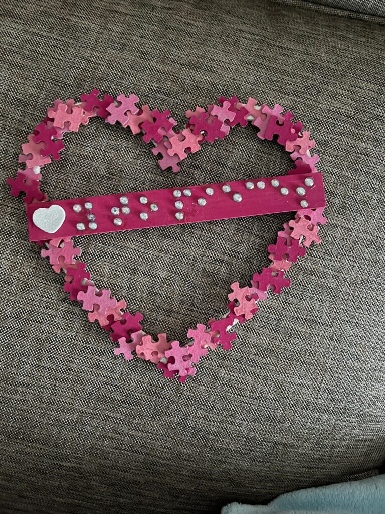 A heart outlined in pink and magenta jigsaw puzzle pieces, with a bright pink wooden bar across the centre spelling a word in raised rhinestone braille dots beside a small white glitter heart.