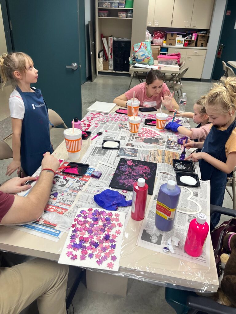 A volunteer helps a group of artists at a table covered in paint trays, cups and loose puzzle pieces in shades of pink, red and purple.