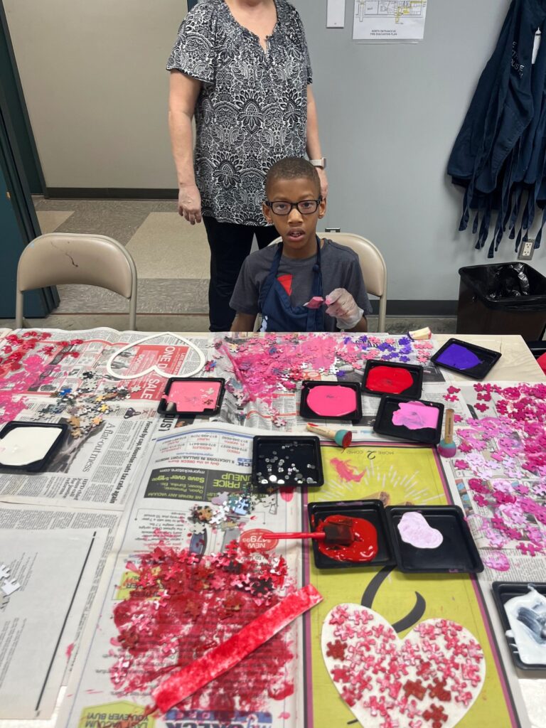 An artist arranges pink and purple puzzle pieces on a heart-shaped card at a table covered in newspaper, paint trays and a bottle of pink acrylic paint.