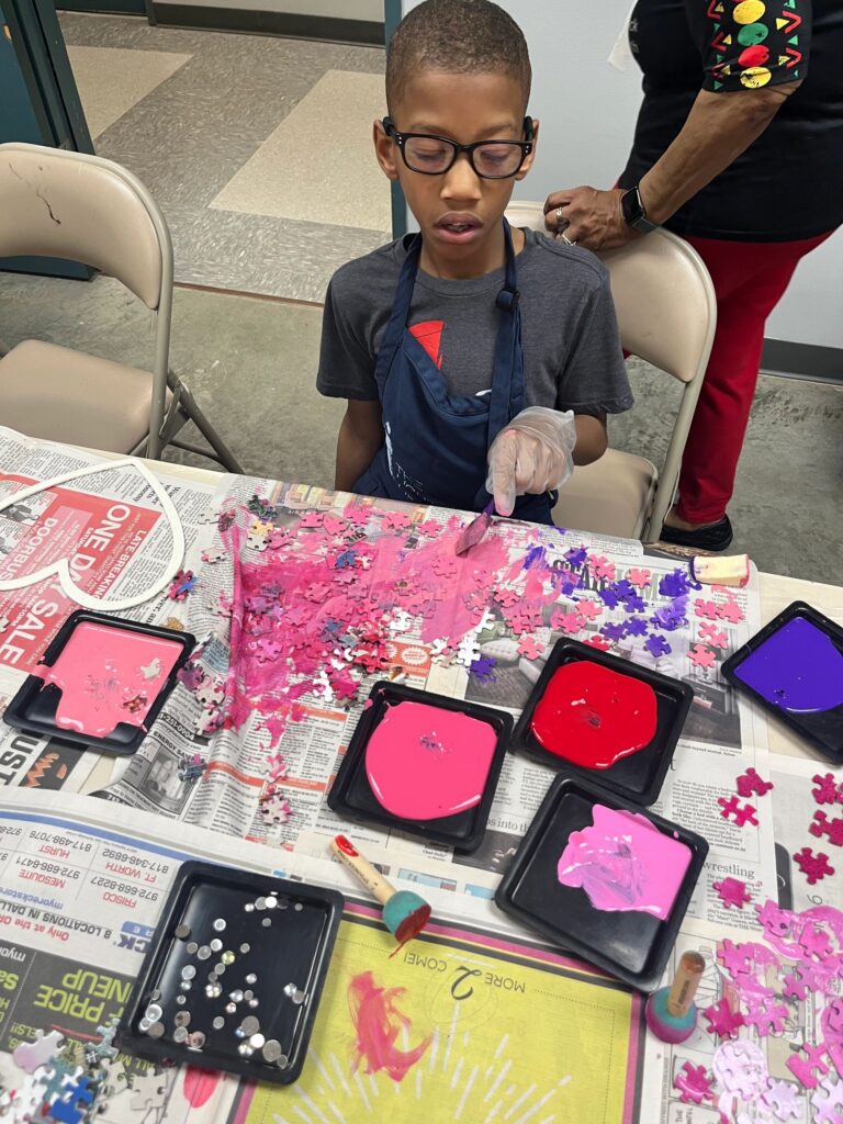 An artist paints red glitter onto a heart-shaped board at a table covered in paint trays, pink paint and scattered puzzle pieces.