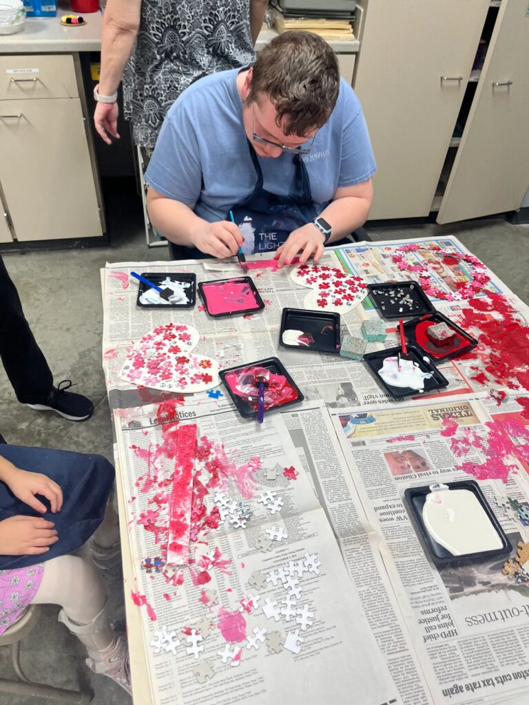 An artist glues loose puzzle pieces onto a heart shape at a table covered in paint trays and pink and white puzzle-piece confetti.