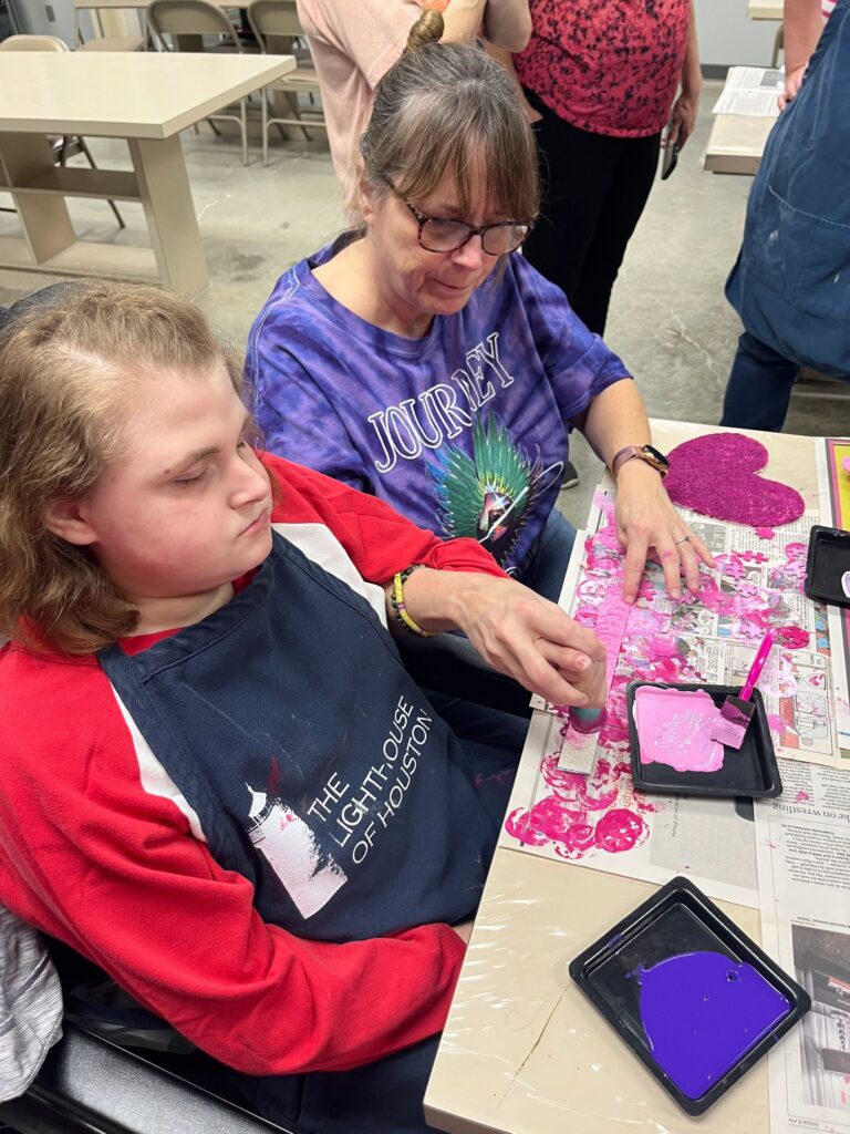 An artist holds up a heart-shaped card covered with a pink and purple jigsaw-puzzle heart, a striped braille bar with a small red heart running across the centre.