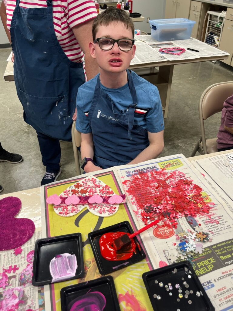 An artist holds a heart-shaped card covered with jigsaw-puzzle pieces, a raised braille bar spelling LOVE running across the middle in raised dots.