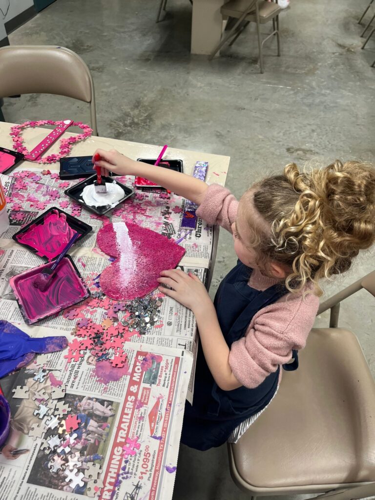 An artist holds a heart made from pink puzzle pieces with a braille bar spelling LOVE across the middle, standing in a hallway near framed watercolour paintings.