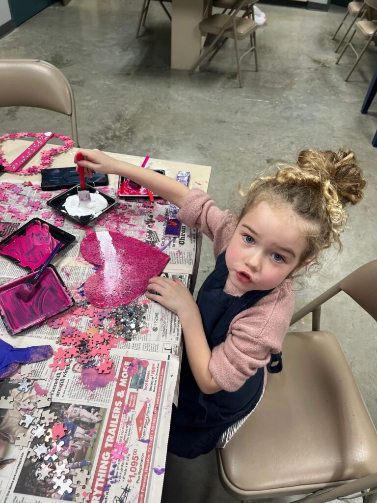An artist holds up a heart made from pink puzzle pieces, with a beaded braille bar spelling LOVE across the centre and a small red heart charm at one end.