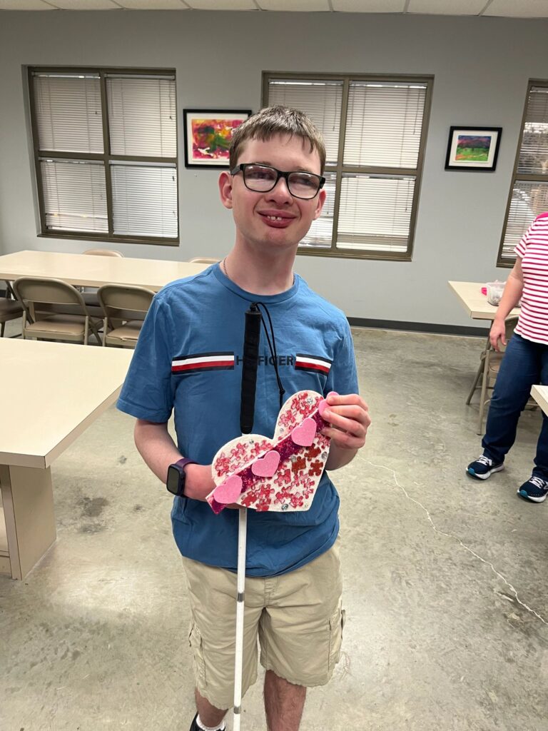 An artist holds a heart made from pink and purple puzzle pieces framing an open centre, with a beaded braille bar and rhinestone trim across the top.