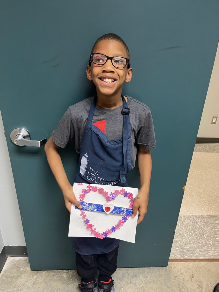 An artist holds a heart made from pink and purple puzzle pieces, framing an open centre with a rhinestone-trimmed braille bar and a small red heart charm.