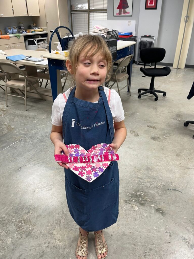 An artist holds up a heart made from pink puzzle pieces layered over a purple and pearl background, decorated with a winged charm at the base.