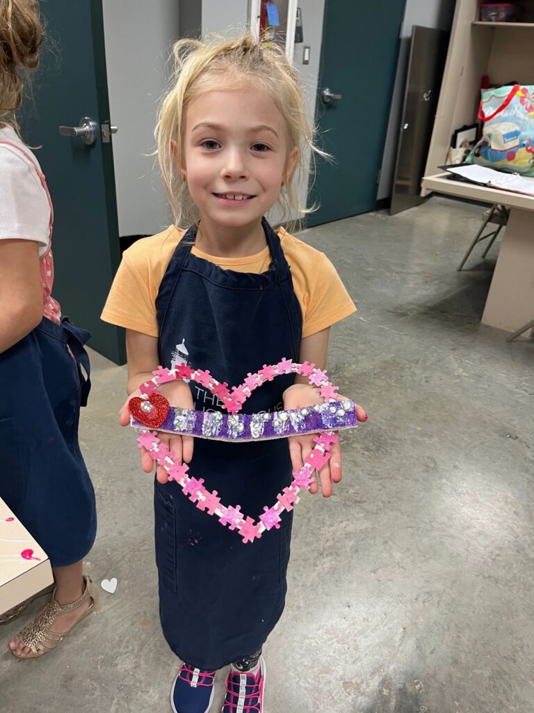 An artist holds a heart-shaped card covered in a dense jigsaw-puzzle pattern in shades of pink and purple, with a rhinestone-trimmed braille bar across the middle.