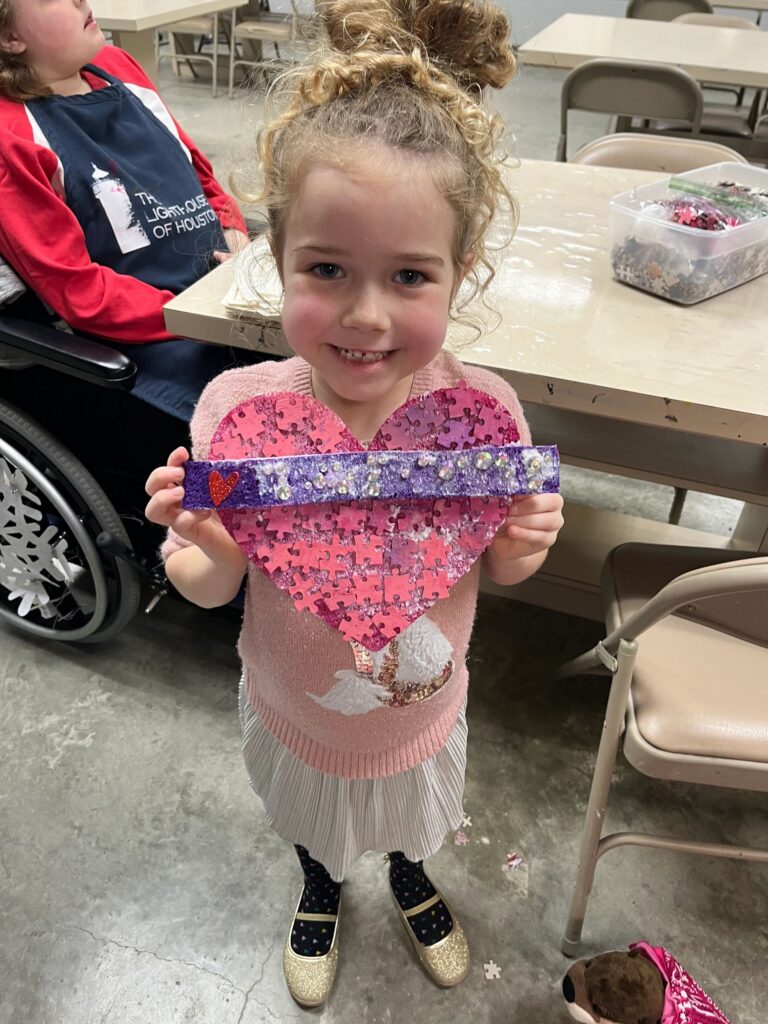 An artist holds up a heart made from pink jigsaw-puzzle pieces with a rhinestone-trimmed purple braille bar and a small red heart charm, in a room with a puzzle-piece bin nearby.